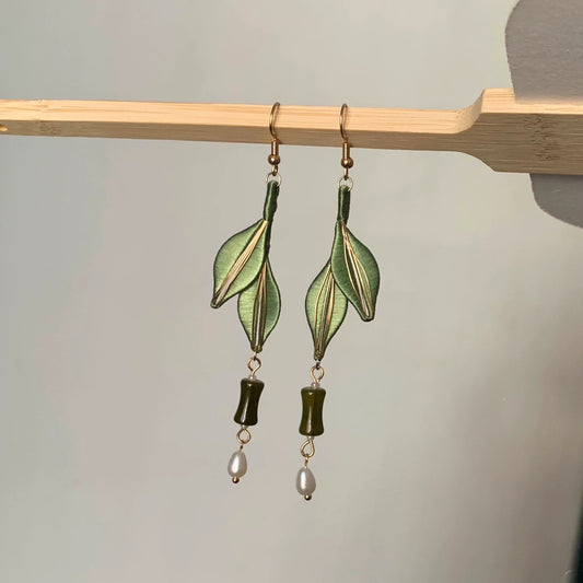 A close-up shot of the handcrafted Chanhua earrings with dark green leaves, a jadeite bead, and a pearl, hanging on a wooden stick.