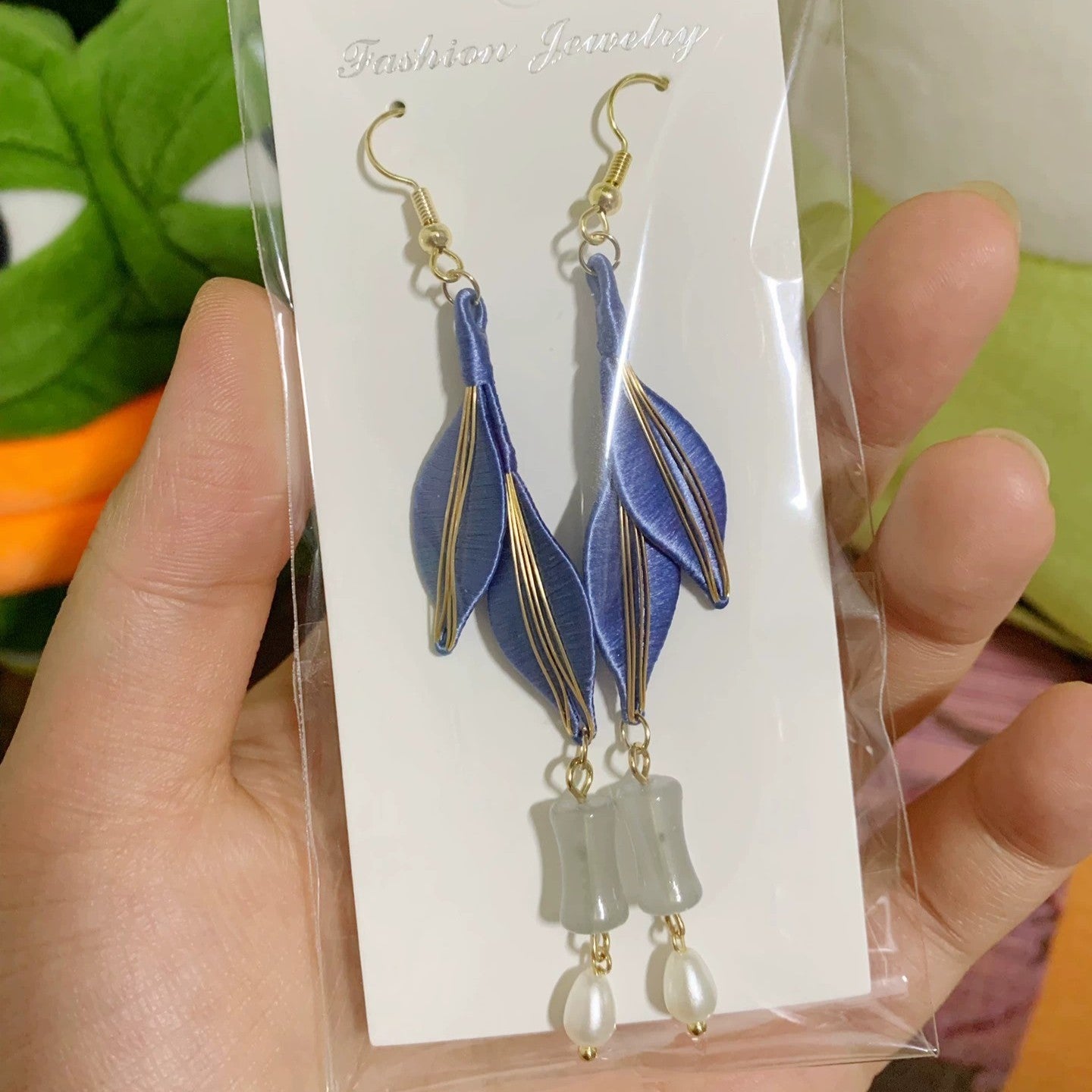 A hand holding a pair of blue-colored Chanhua earrings in a clear plastic package, showing the intricate design and materials.