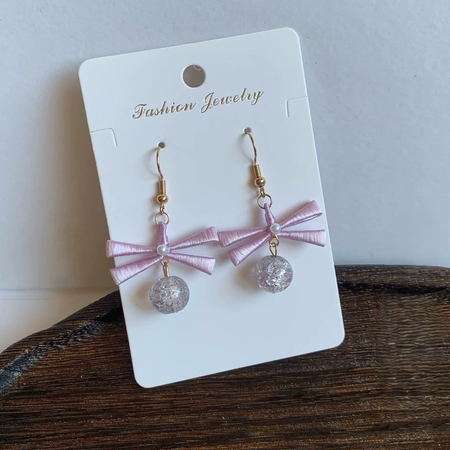 A pair of handmade Chanhua earrings with a light pink ribbon bow and a clear, shimmering beaded drop.