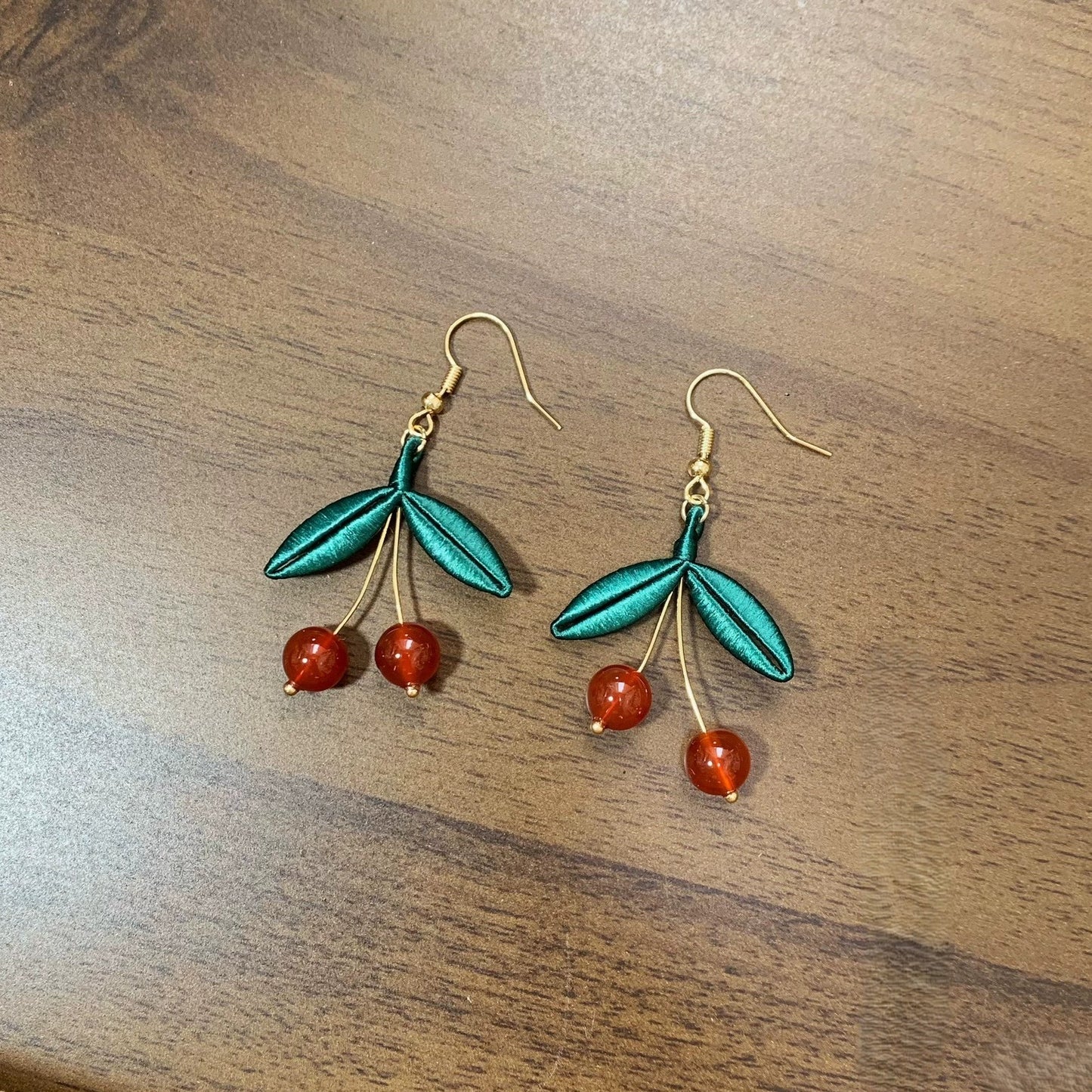 A pair of handmade Chanhua earrings featuring a red cherry design with green silk leaves on a wooden background.