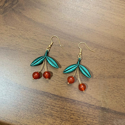 A pair of handmade Chanhua earrings featuring a red cherry design with green silk leaves on a wooden background.