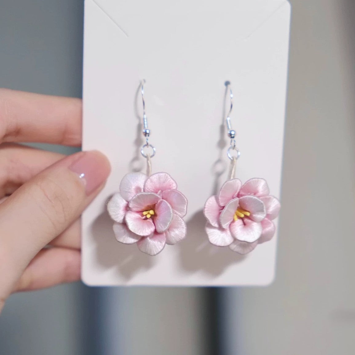 Hand holding a pair of handmade pink camellia flower dangle earrings with 925 sterling silver hooks on a white display card.