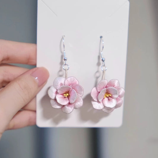 Hand holding a pair of handmade pink camellia flower dangle earrings with 925 sterling silver hooks on a white display card.