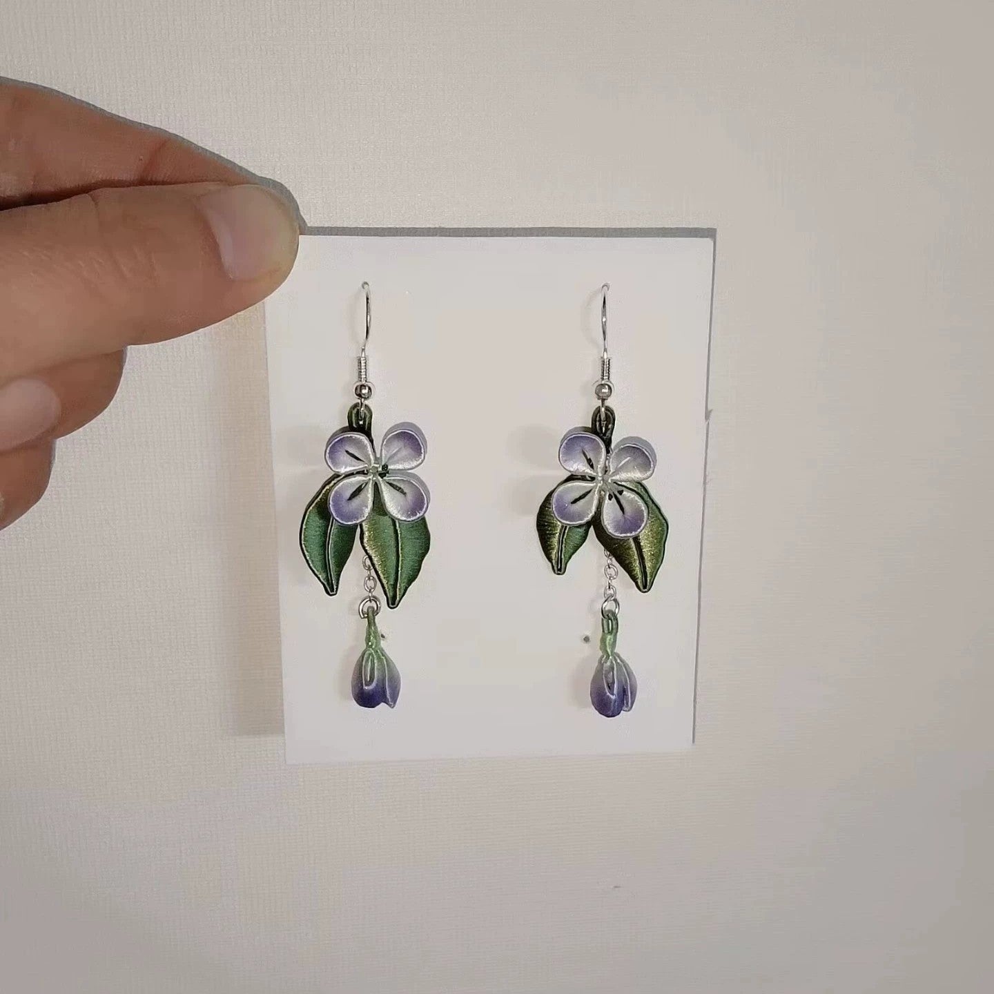 Handcrafted Chanhua flower earrings with purple petals and green leaves, held against a white background.