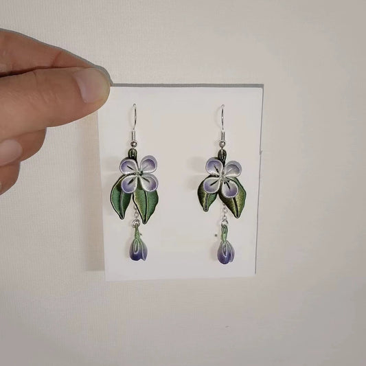 Handcrafted Chanhua flower earrings with purple petals and green leaves, held against a white background.