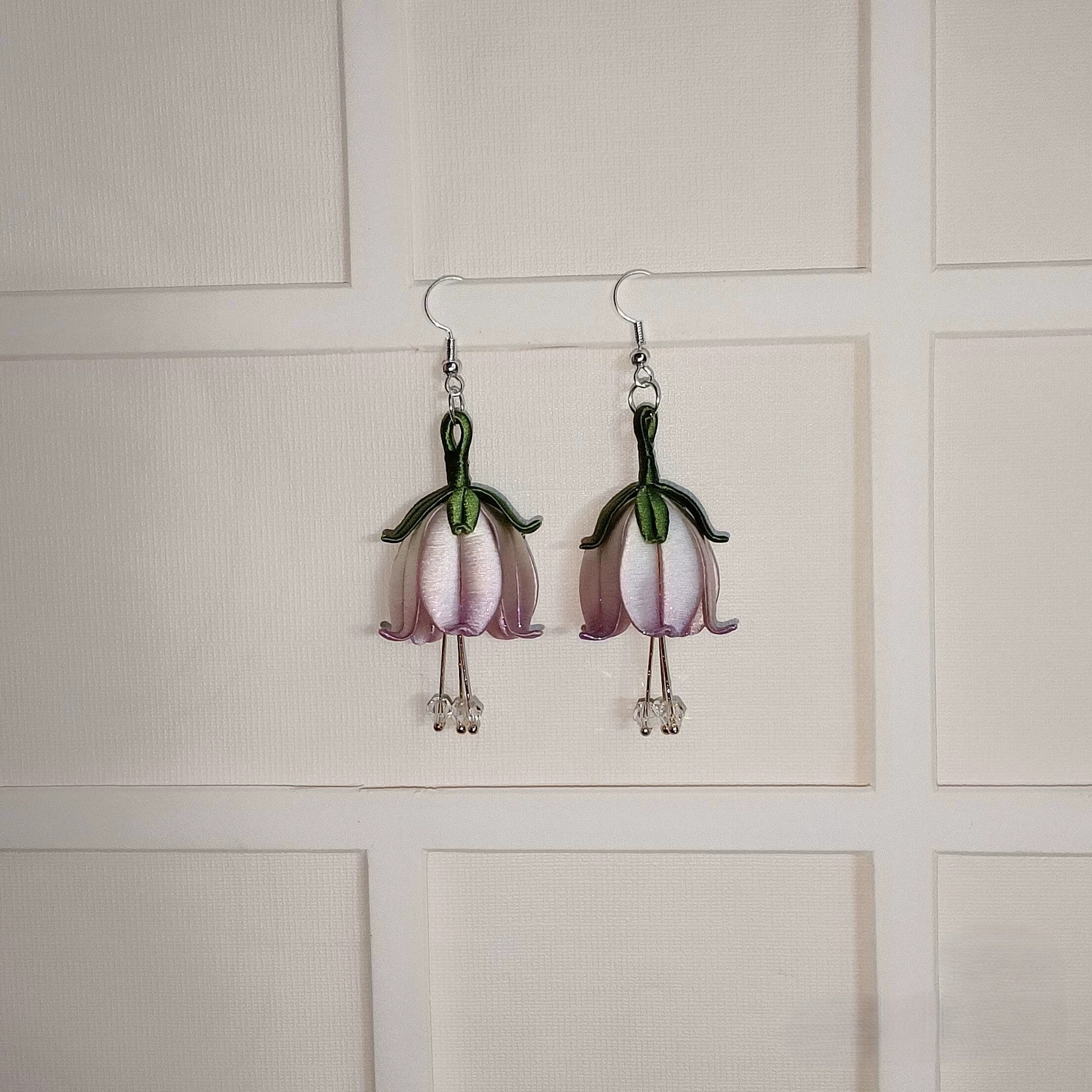 A pair of handmade Chanhua flower dangle earrings on a white textured background. The earrings feature light purple petals and a green stem, with small crystal beads at the bottom.