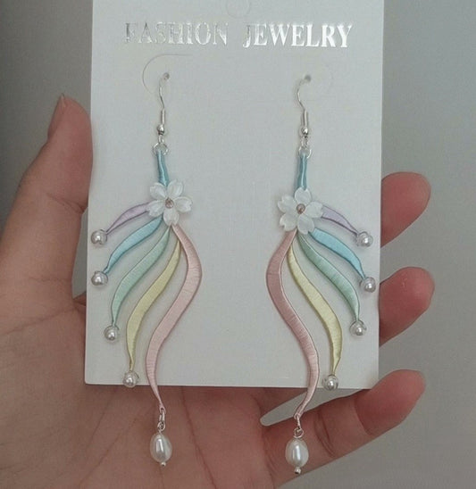 A pair of handmade Chanhua earrings featuring pastel silk tassels and a white flower, held against a white background.