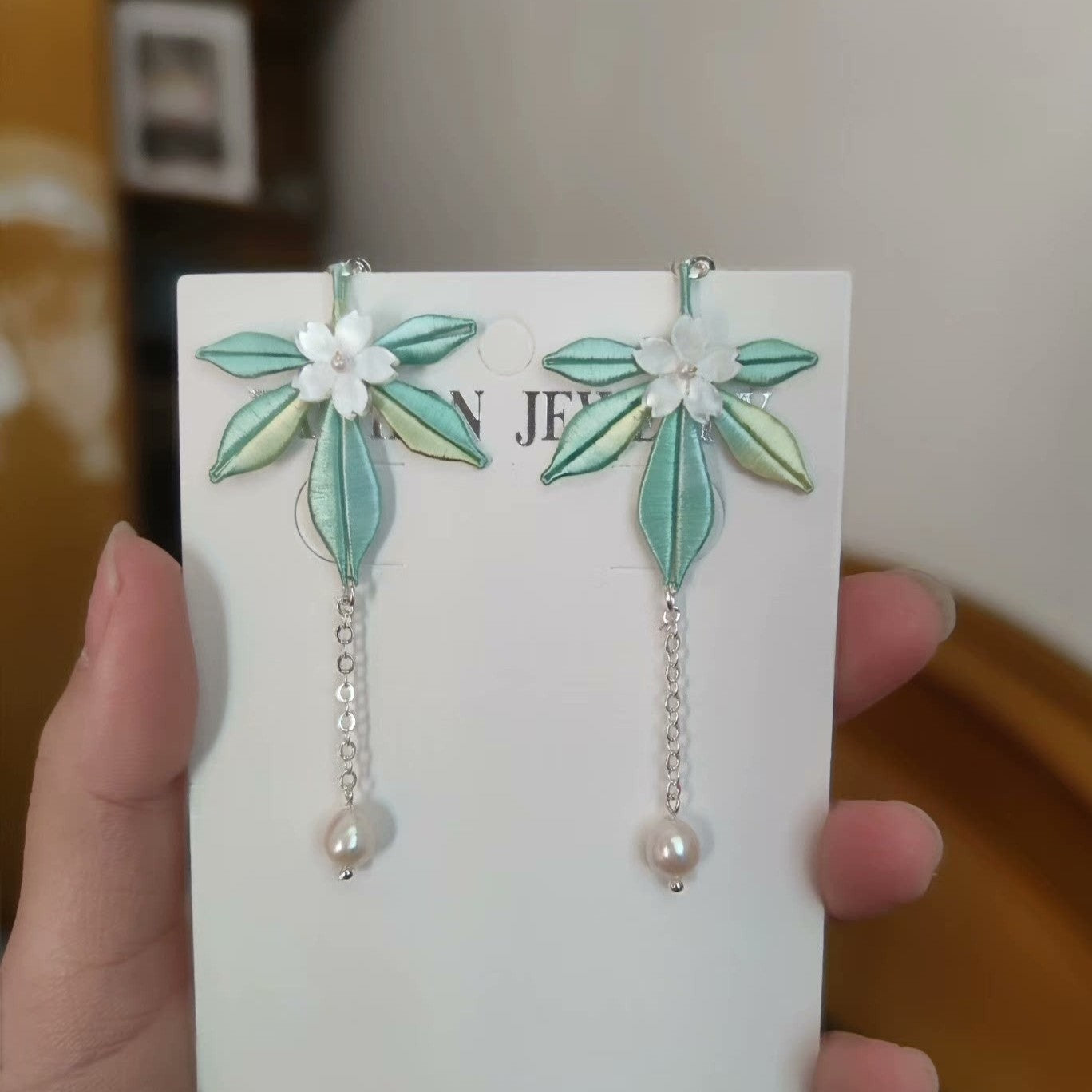 A hand holding a pair of light green handmade Chanhua earrings featuring a small white flower detail.