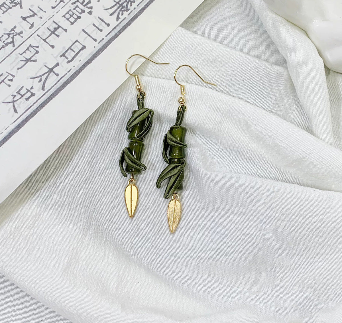 A close-up shot of the handmade bamboo and leaf dangle earrings on a white cloth background with an open book.