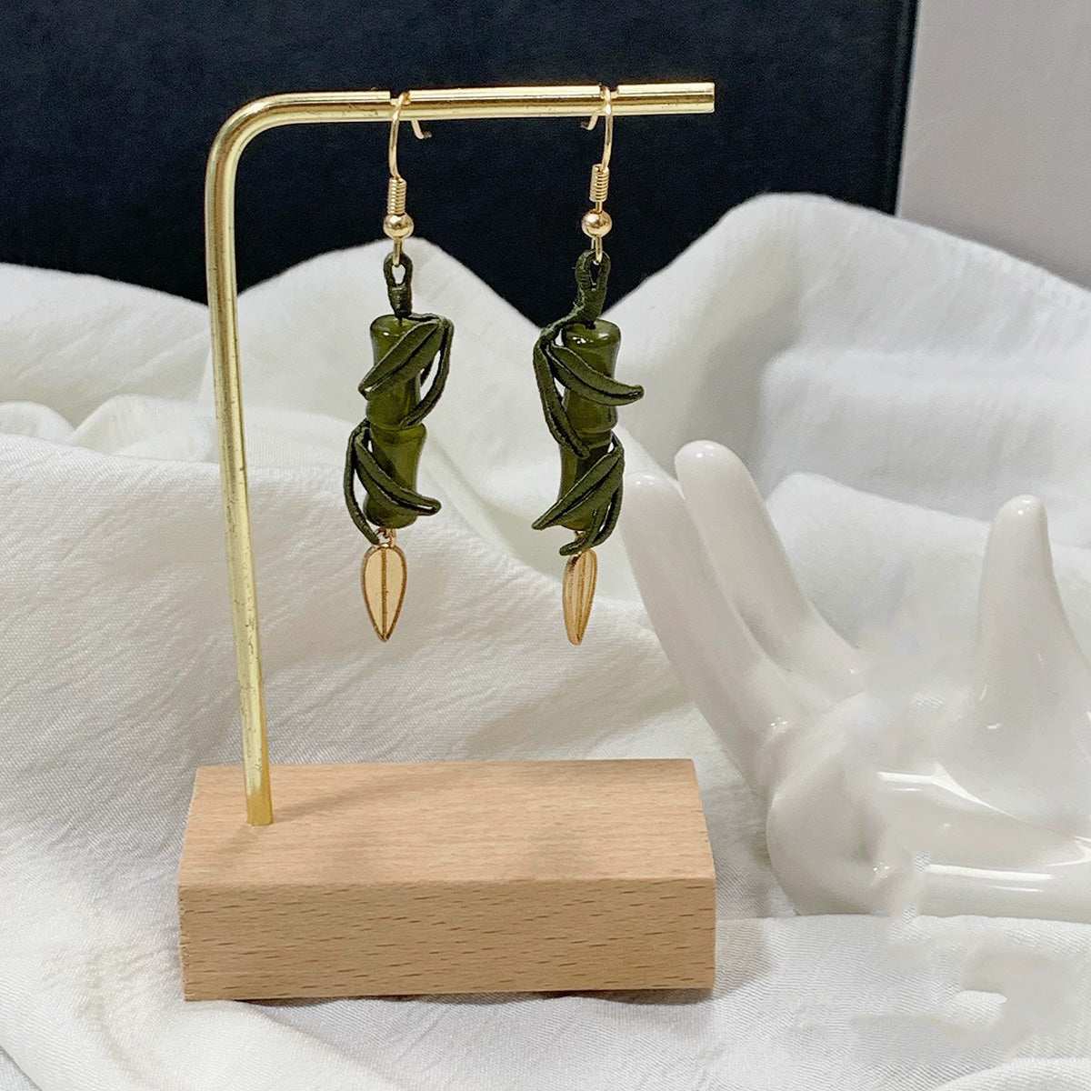 A pair of handmade 925 sterling silver hook earrings with green bamboo and golden leaf charms, displayed on a wooden stand.