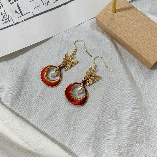 A pair of handmade Chanhua earrings with a golden butterfly and a red embroidered circle.