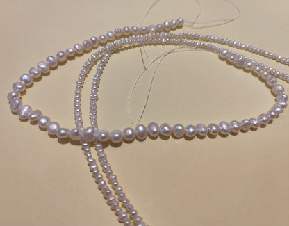 Two long strands of small, natural freshwater baroque pearls laid out on a crafting mat.
