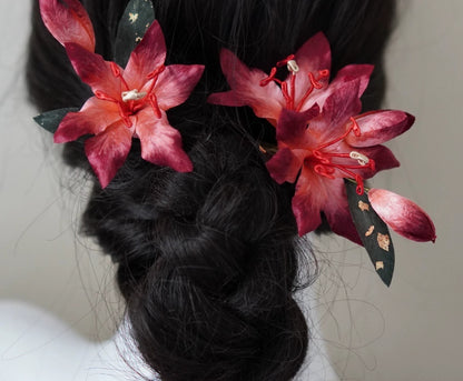 Close-up of two handcrafted crimson Mulberry Silk Ronghua Lily hair pins securing a dark, braided hairstyle, showing the floral details.