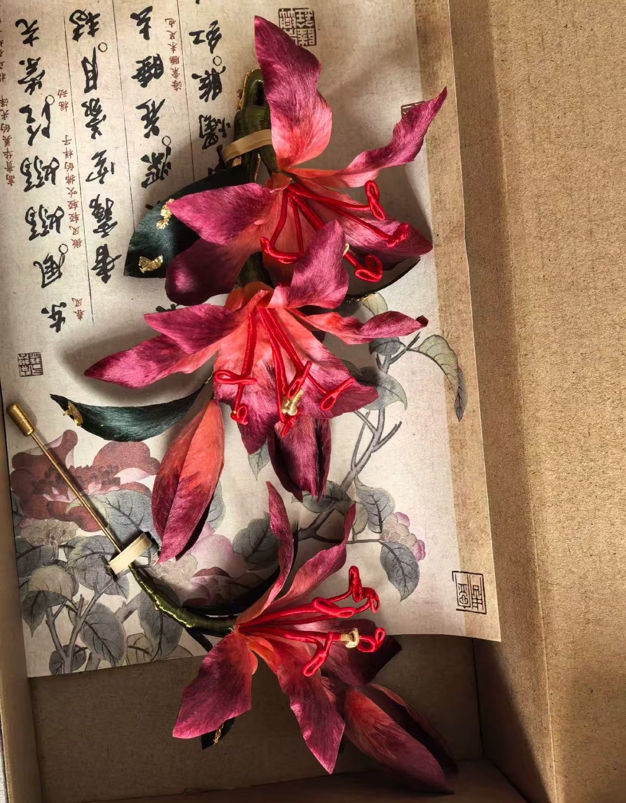 Handmade deep red Mulberry Silk Ronghua Lily hair pin and brooch set displayed on traditional artwork, highlighting the rich color and craftsmanship.