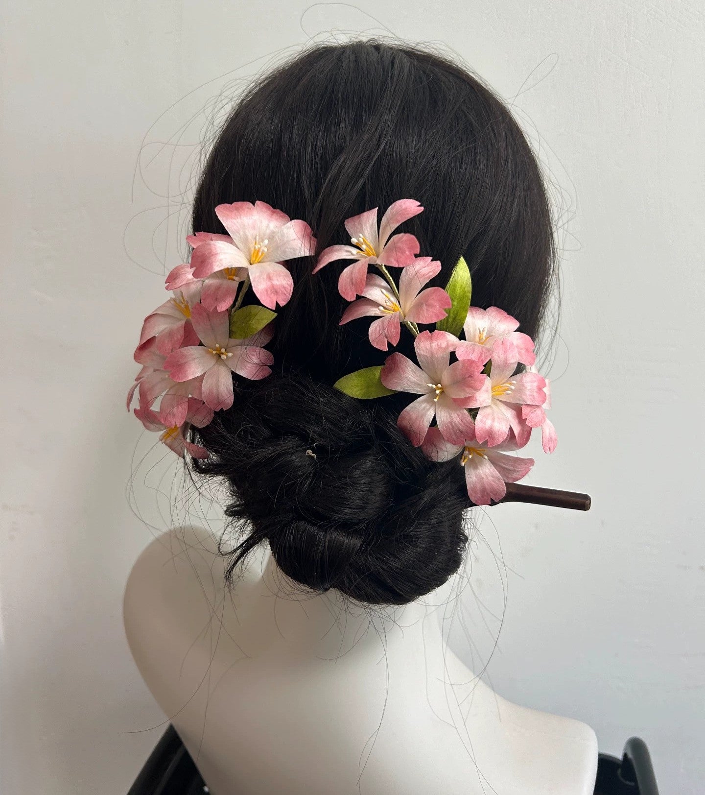 A dark-haired mannequin wearing a large, handcrafted Ronghua cherry blossom hair stick, showing the pink mulberry silk flowers clustered beautifully on a low hair bun.