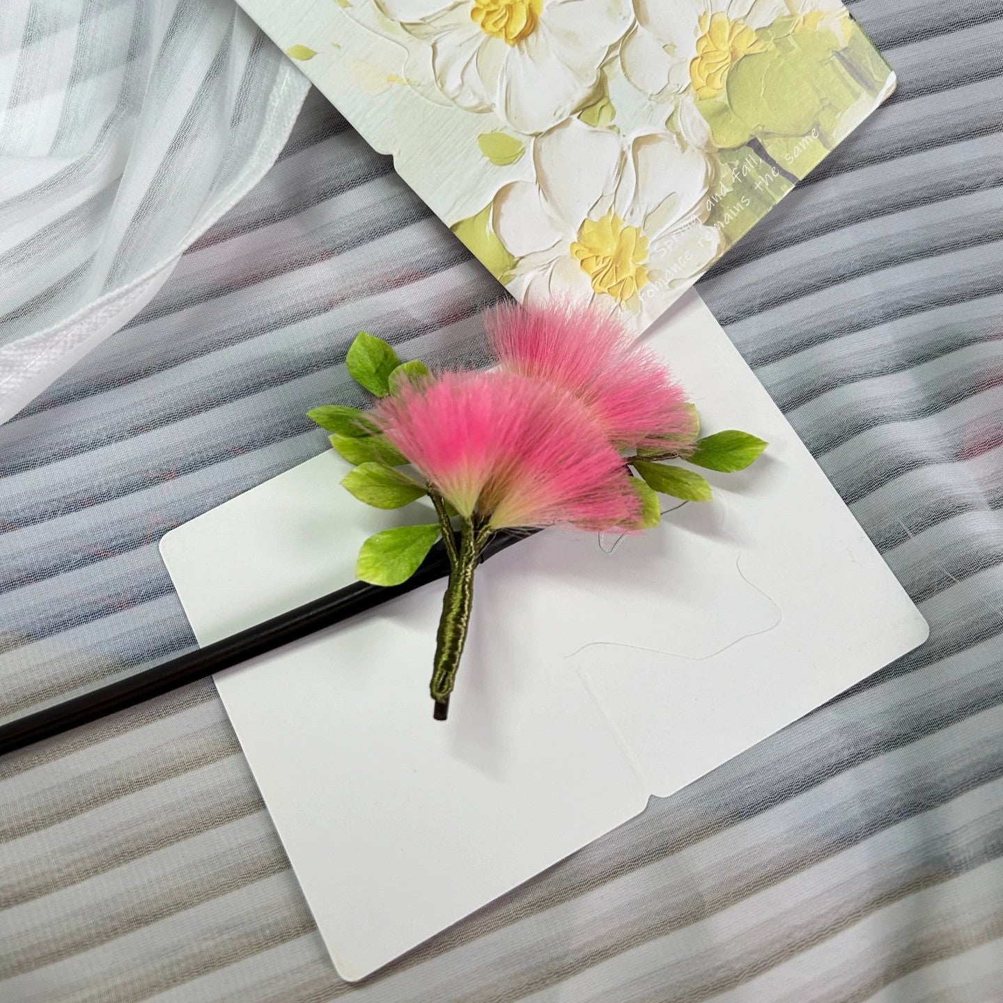 Handheld close-up of handmade pink Ronghua silk Albizia flower hair stick with fluffy mimosa blooms and green leaves