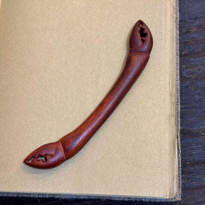 Natural African Padauk hair stick with crab claw design 13.5cm on open book background.